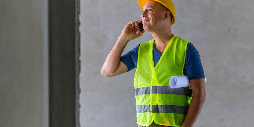 Construction contractor in a hard hat and safety vest on a phone call at a job site, holding rolled plans