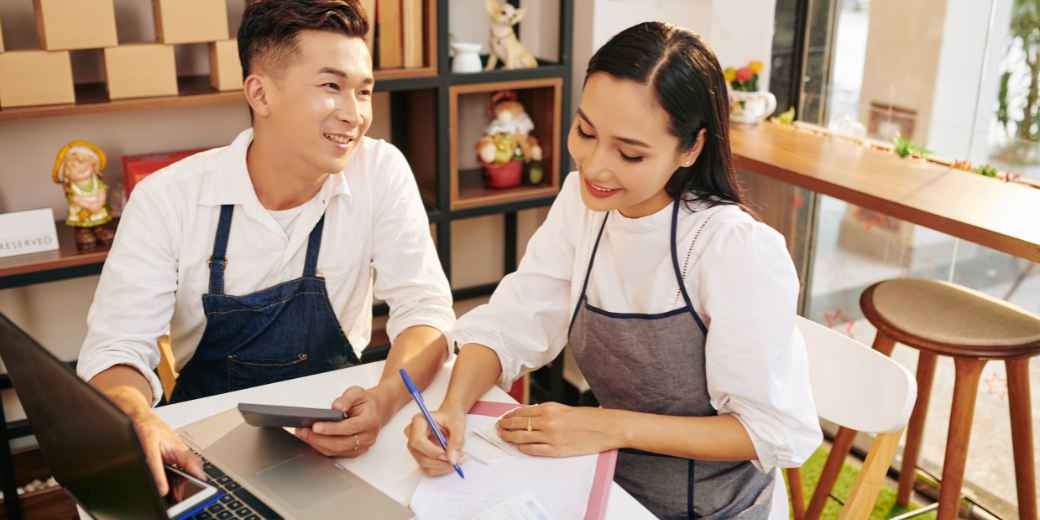 Two shop owners in aprons going over paperwork with a laptop and calculator