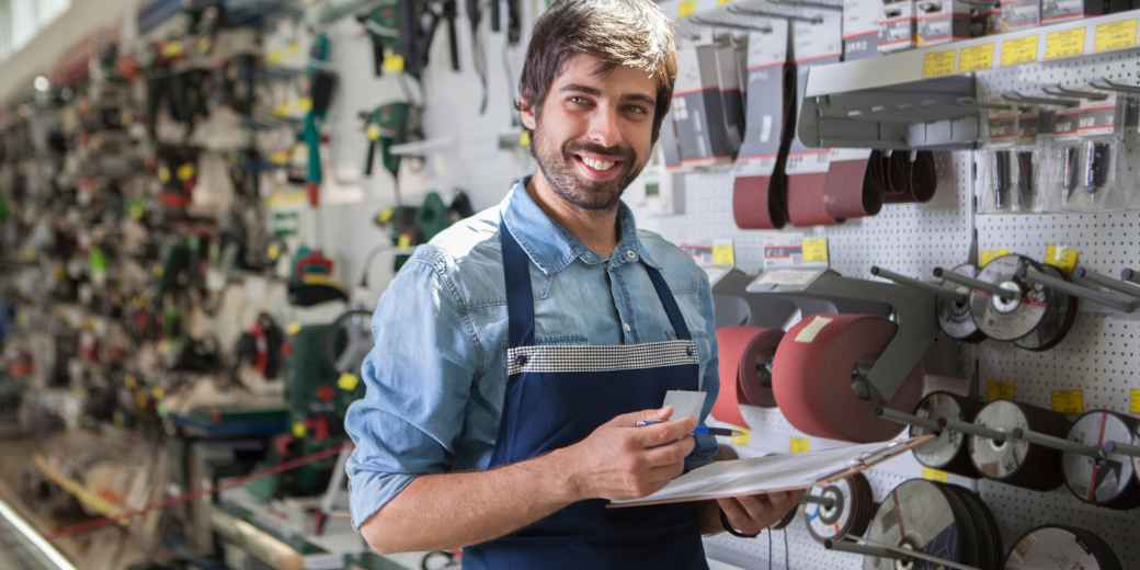 Hardware store owner in an apron holding a clipboard in front of a wall of tools