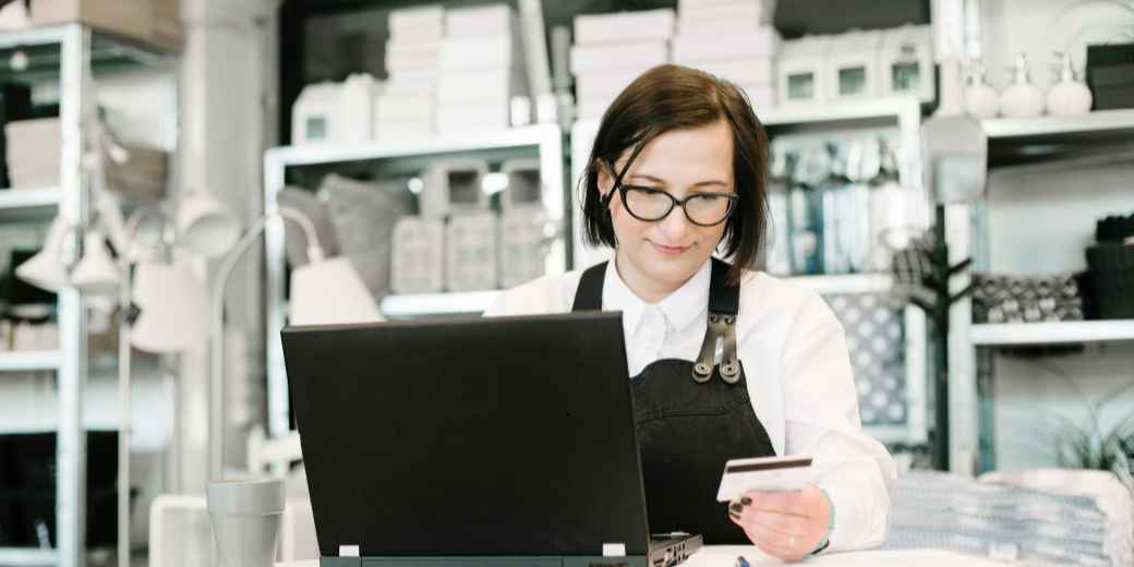Home goods shop owner in glasses and an apron at her laptop behind the counter, holding a payment card