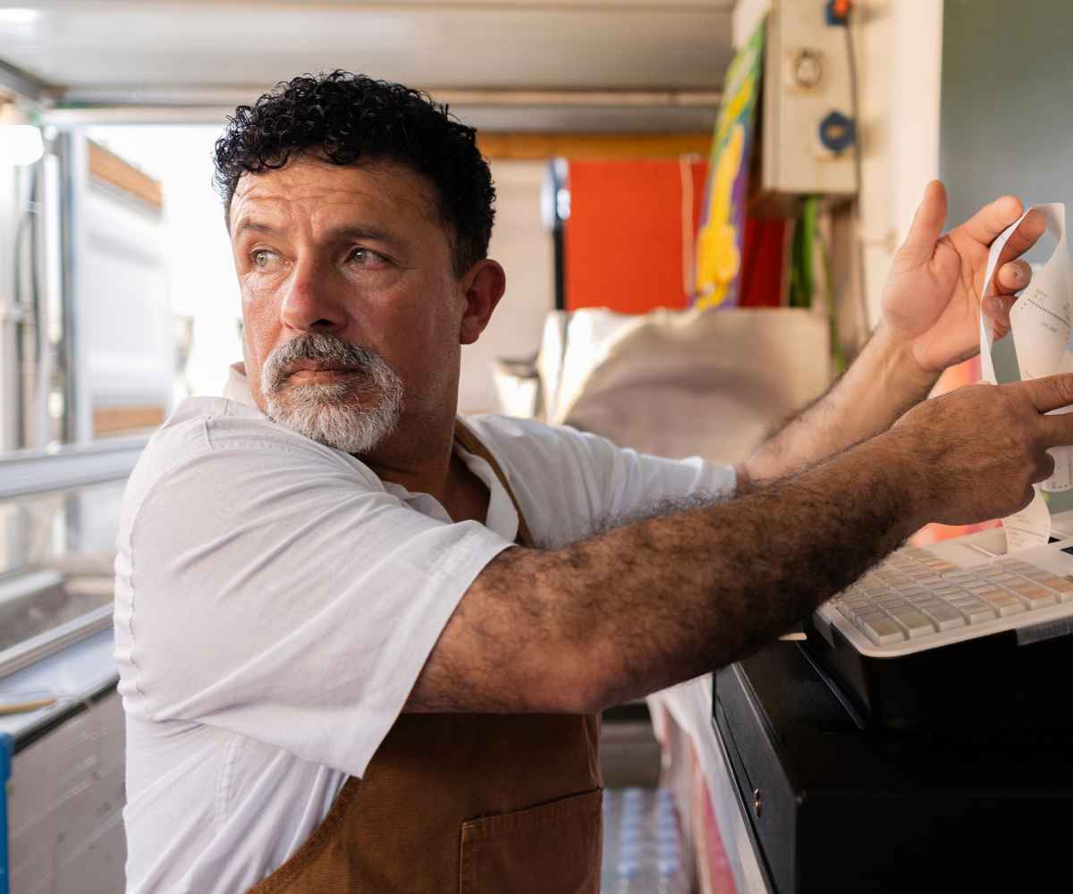 Food truck owner in a brown apron pulling an order ticket from the printer inside his truck, looking off in thought