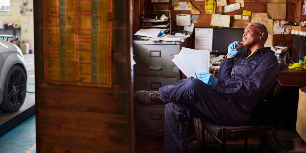 Auto repair shop owner in work coveralls and gloves sitting in a garage office, holding paperwork and looking off in thought