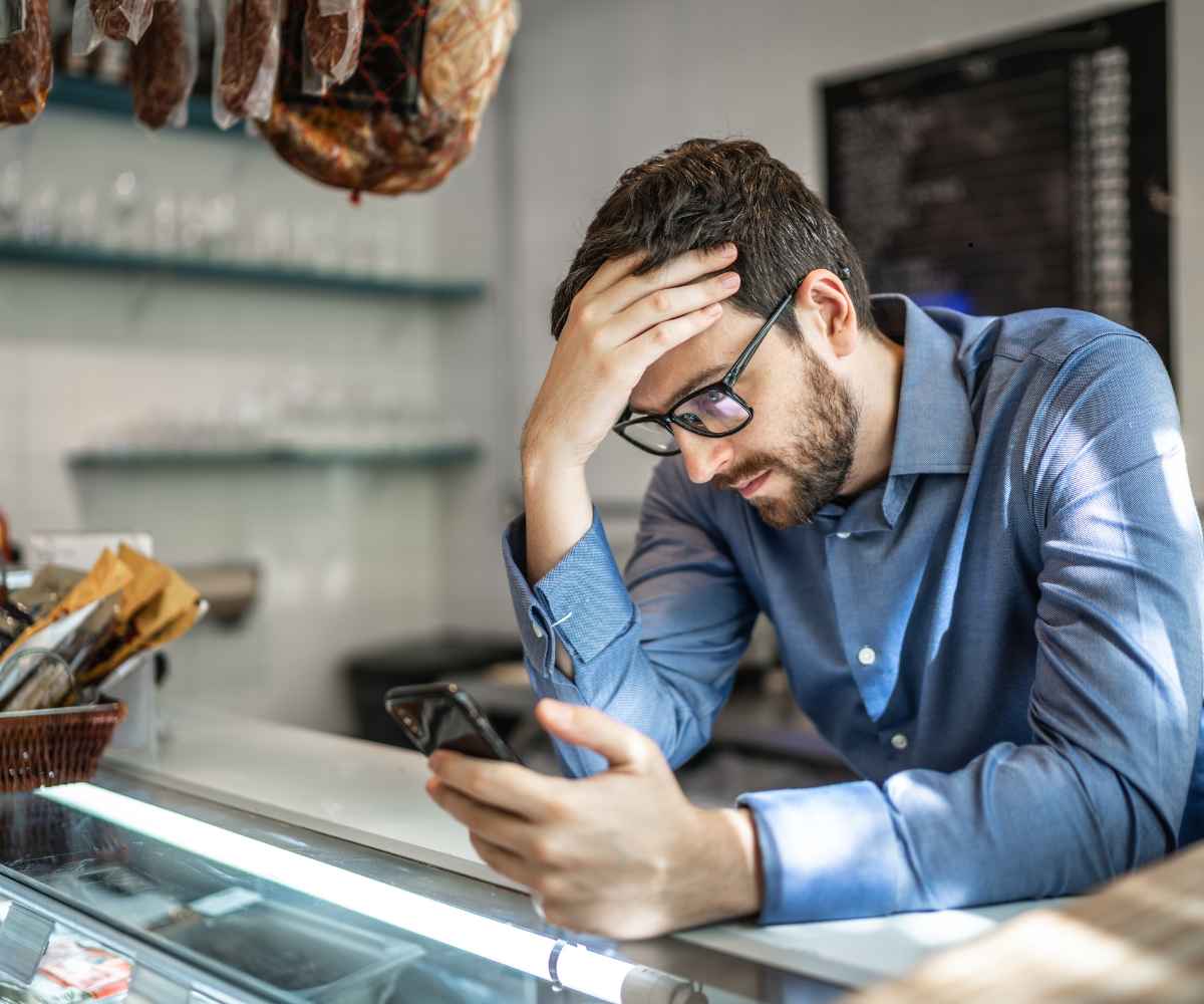Deli owner behind his counter with his hand on his forehead, reading his phone, worried about the personal guarantee on his merchant cash advance