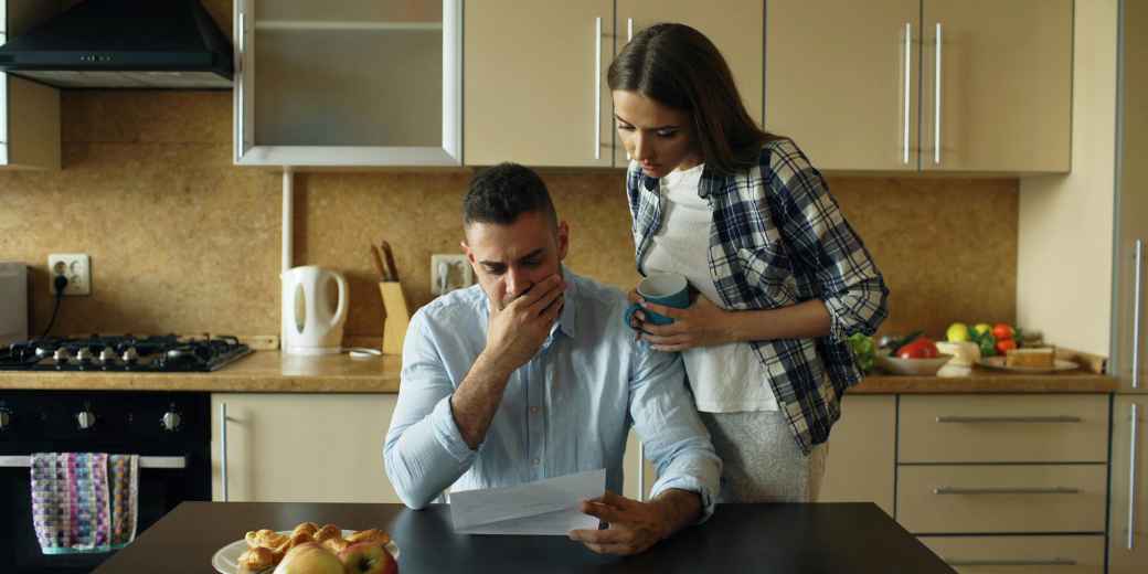 Man at his kitchen table reading a letter with his hand over his mouth while his partner looks on