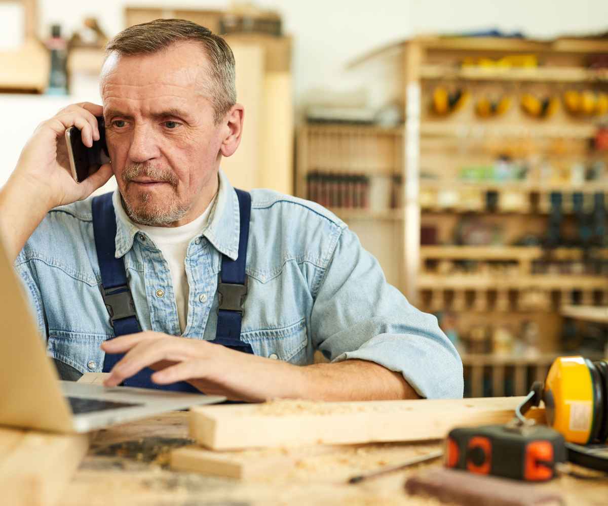 Woodshop owner in overalls on a phone call at his laptop, with lumber and tools on the workbench