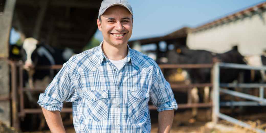 Dairy farmer in a ball cap and plaid shirt smiling in front of his barn