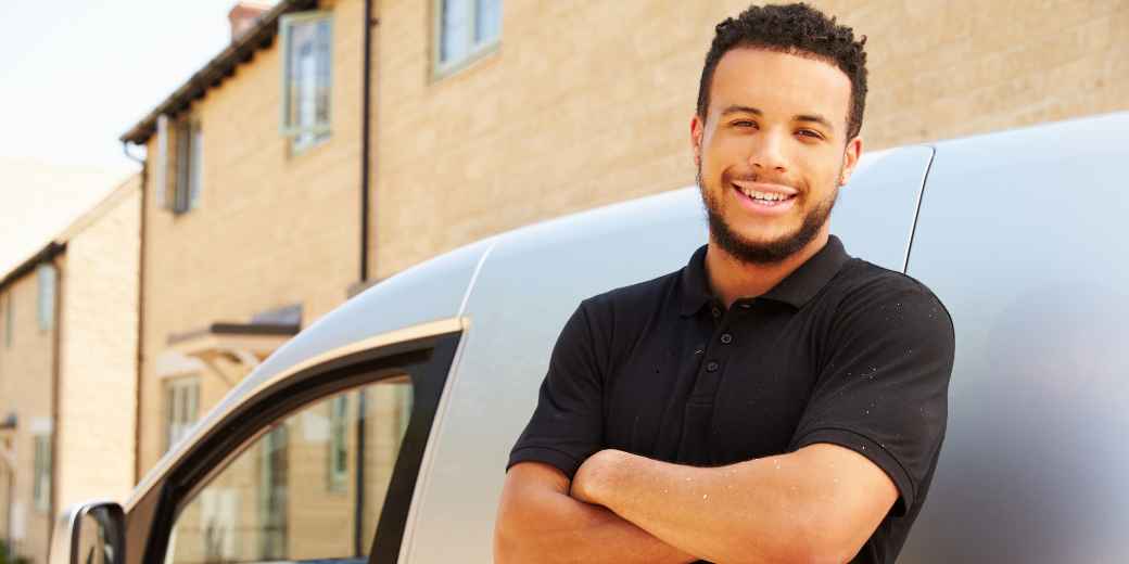 A smiling tradesman with his arms crossed, leaning on his work van outside a stone building
