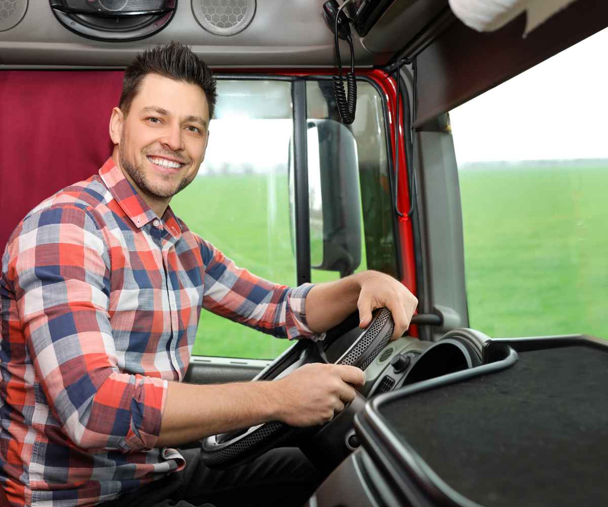 A smiling truck owner-operator in a plaid shirt sitting at the wheel of his truck cab