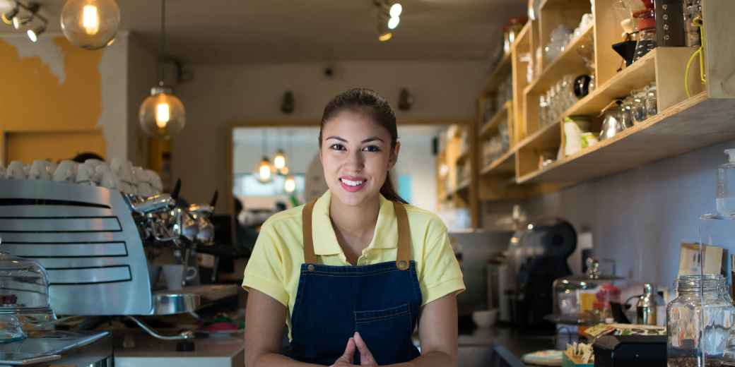 A smiling restaurant owner in an apron leaning on her counter, with the coffee bar behind her
