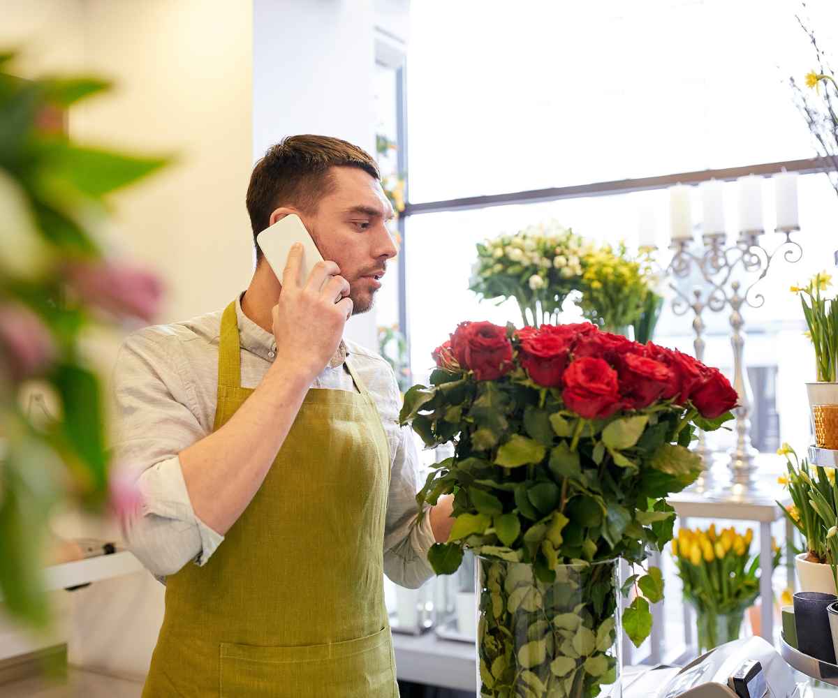 Florist in a green apron on a phone call while arranging red roses in his flower shop