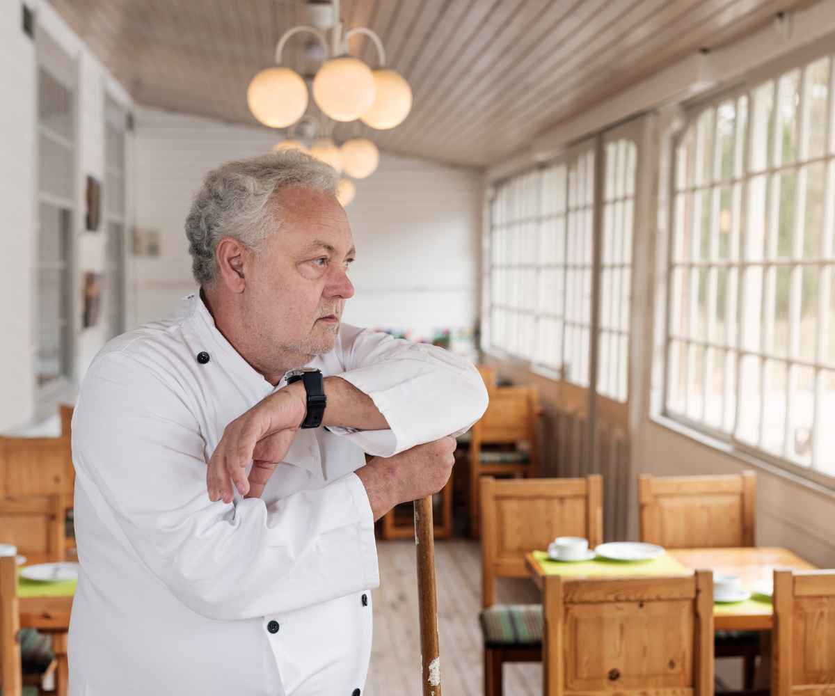 Restaurant owner in a white chef's coat leaning on a chair in his empty dining room, looking out the window and weighing his small business debt