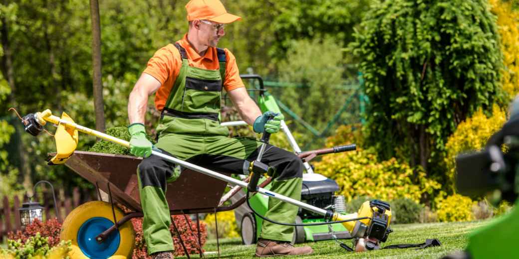 Landscaper in an orange cap and green overalls resting on a wheelbarrow with a string trimmer across his lap